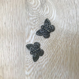 May include: Two decorative metal butterfly charms with intricate cut-out designs. The butterflies are a dark, antique bronze color and are lying on a light-colored wooden surface. These charms could be used for jewelry making or craft projects.