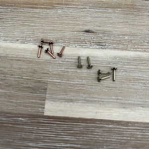 May include: A collection of small metal rivets in various colors, including copper and brass tones, are scattered on a light brown wooden surface. The rivets have a T-shaped head and a cylindrical body, suitable for leathercraft or similar projects.