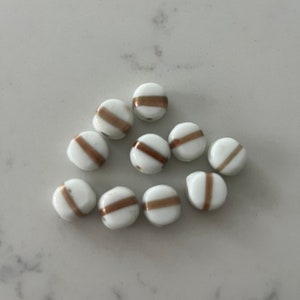 May include: A group of white glass beads with a single gold stripe around the centre of each bead.