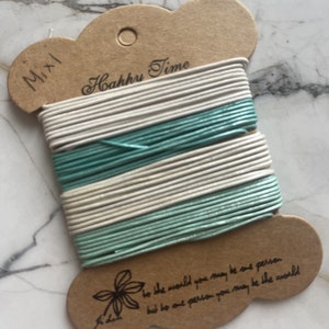 May include: A spool of cord in shades of white and teal, mounted on a brown card. The card has the text "Mixl" and "Hahhu Time" printed on it. The bottom of the card has the text "to the world you may be one person but to one person you may be the world".