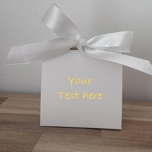May include: A white square gift box tied with a white satin ribbon bow. The box has the words "Your text here" printed in yellow. The box is on a wooden surface.