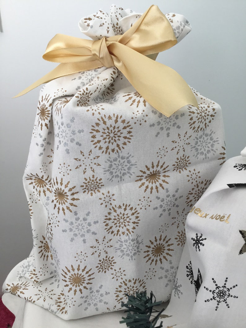 Puede incluir: Una bolsa de regalo de tela blanca con una cinta dorada y un patr&oacute;n de copos de nieve dorados y plateados. La bolsa est&aacute; atada con un lazo y tiene un ambiente festivo.