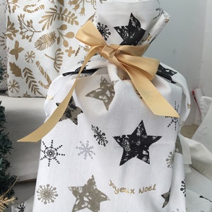 Puede incluir: Una bolsa de regalo blanca con una cinta dorada y un patr&oacute;n de estrellas negras y doradas. La bolsa tiene el texto "Joyeux Noel" impreso.