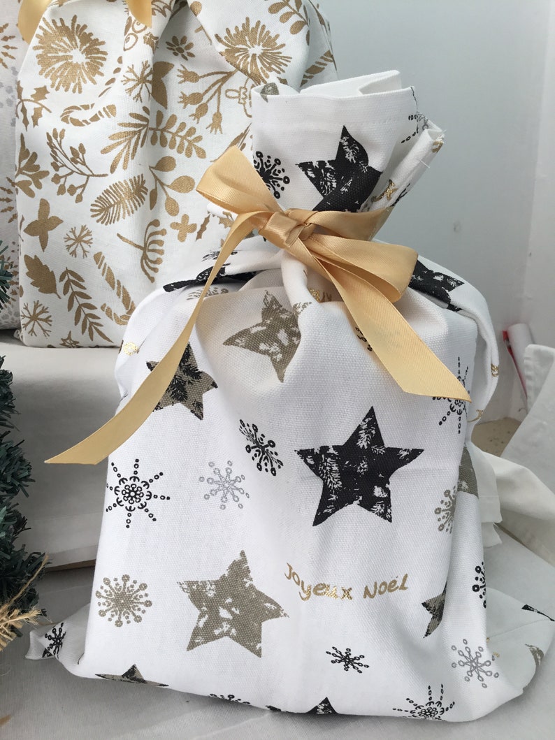Puede incluir: Una bolsa de regalo blanca con una cinta dorada y un patr&oacute;n de estrellas negras y doradas. La bolsa tiene el texto "Joyeux Noel" impreso.