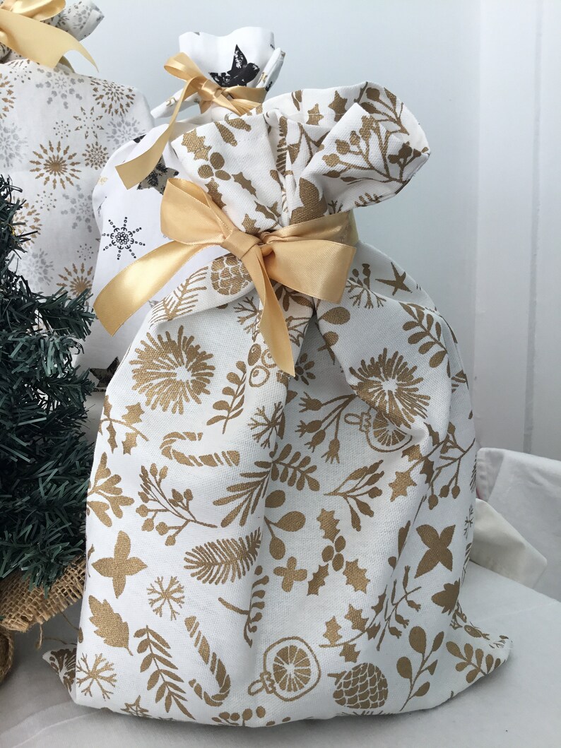 Puede incluir: Una bolsa de regalo de tela blanca con un patr&oacute;n floral dorado y una cinta dorada atada alrededor de la parte superior. La bolsa est&aacute; llena de un regalo.