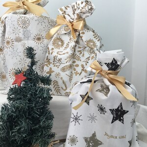 Puede incluir: Tres bolsas de regalo de tela blanca con cinta dorada y diferentes patrones. Las bolsas est&aacute;n decoradas con copos de nieve dorados, estrellas y dise&ntilde;os florales. La bolsa de la derecha tiene el texto "Joyeux Noel" en letras doradas.