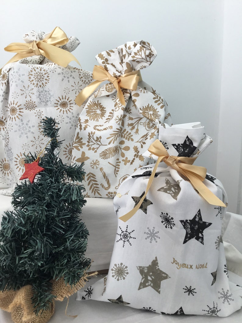 Puede incluir: Tres bolsas de regalo de tela blanca con cinta dorada y diferentes patrones. Las bolsas est&aacute;n decoradas con copos de nieve dorados, estrellas y dise&ntilde;os florales. La bolsa de la derecha tiene el texto "Joyeux Noel" en letras doradas.
