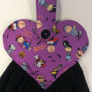 Peut inclure: Serviette de cuisine en forme de c&oelig;ur violette avec une serviette noire attach&eacute;e. Le tissu violet pr&eacute;sente un motif de personnages de Peanuts habill&eacute;s en costumes d'Halloween, des chauves-souris et les mots "Boo!" et "Trick or Treat".