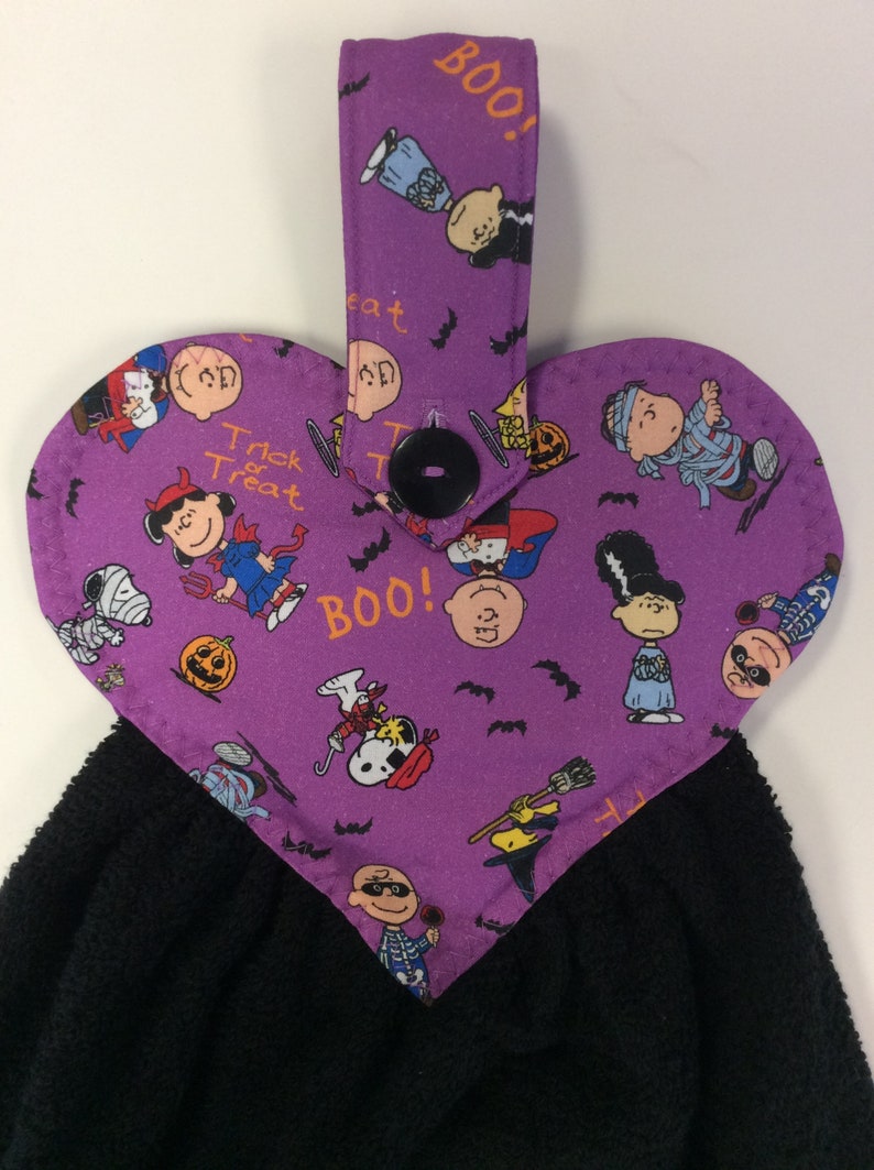 Peut inclure: Serviette de cuisine en forme de c&oelig;ur violette avec une serviette noire attach&eacute;e. Le tissu violet pr&eacute;sente un motif de personnages de Peanuts habill&eacute;s en costumes d'Halloween, des chauves-souris et les mots "Boo!" et "Trick or Treat".