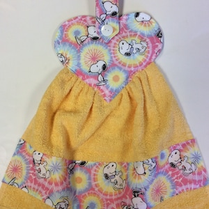 May include: Yellow kitchen towel with a heart-shaped hanging loop. The loop is made of tie-dye fabric with a white Snoopy print. The towel has a tie-dye border with a white Snoopy print.