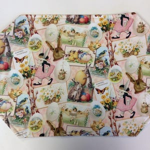 May include: A white fabric placemat with a pink background featuring a repeating pattern of Easter-themed images. The images include bunnies, chicks, eggs, flowers, and birds. The placemat has a scalloped edge.