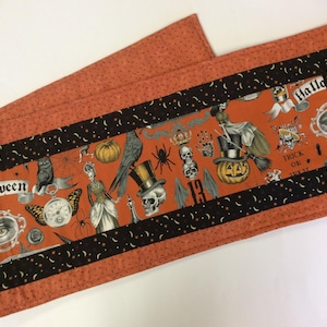 May include: Halloween-themed table runner with an orange background and a black border. The center features Halloween-related illustrations, including pumpkins, skulls, and the word "Halloween". The runner is approximately 12 inches wide and 36 inches long.