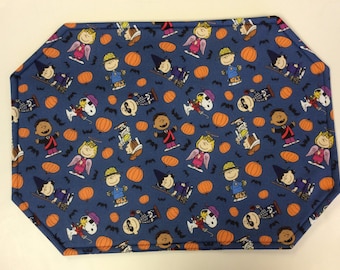 May include: A blue fabric placemat with a Halloween theme featuring Peanuts characters like Charlie Brown, Snoopy, and Linus. The placemat is covered in a repeating pattern of pumpkins, bats, and Peanuts characters dressed in Halloween costumes.