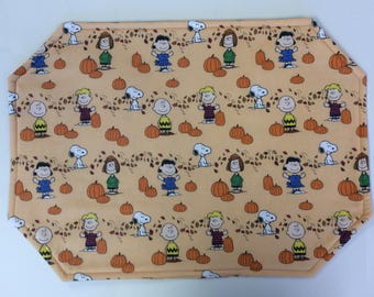 Peut inclure: Un set de table rectangulaire en tissu sur fond orange clair, orné d'un motif répétitif représentant les personnages des Peanuts comme Snoopy, Charlie Brown, Lucy et Linus, entourés de citrouilles orange et de feuilles d'automne brunes.
