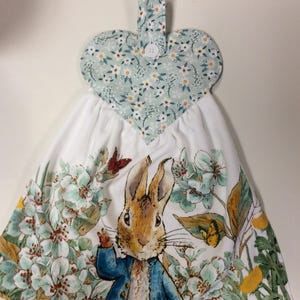 May include: A white tea towel with a Peter Rabbit design featuring a rabbit in a blue jacket surrounded by flowers. The towel has a heart-shaped hanging loop with a floral pattern.