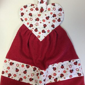 May include: Red and white kitchen towel with a heart-shaped hanging loop. The towel features a ladybug print with red ladybugs, white dots, green leaves, and white flowers on a white background.