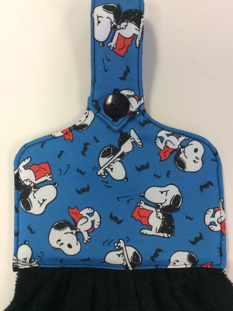 Peut inclure: Une serviette de cuisine bleue avec un bouton noir et une boucle de suspension. La serviette pr&eacute;sente un motif r&eacute;p&eacute;t&eacute; de Snoopy d&eacute;guis&eacute; en vampire avec une cape rouge et des chauves-souris.