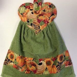 May include: Green kitchen towel with a heart-shaped hanging loop. The loop is decorated with a fall-themed fabric featuring pumpkins, sunflowers, and corn. The towel has a green border and a bottom band of the same fall-themed fabric.