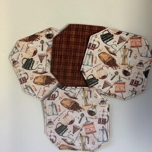 May include: A set of five octagonal fabric coasters. Four coasters feature a coffee-themed pattern with coffee beans, coffee pots, and cups. The center coaster has a brown plaid pattern. The coasters are arranged on a white surface.