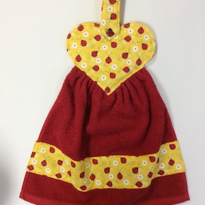 May include: Red and yellow kitchen towel with a heart-shaped hanging loop. The towel is decorated with a ladybug and flower pattern.