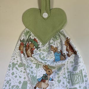 May include: A heart-shaped kitchen towel holder with a green polka-dot design and a white button. The towel features a white background with a pattern of green leaves and illustrations of rabbits in clothing.