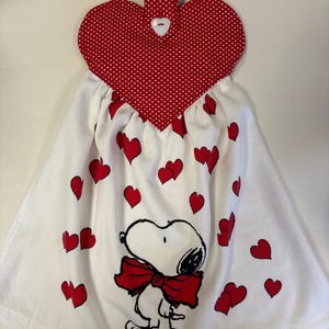 May include: White kitchen towel with red heart accents and a Snoopy graphic wearing a red bow tie. The top features a red heart-shaped fabric with white polka dots and a white button. A red fabric loop is at the top for hanging.