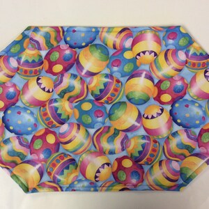 May include: A blue fabric placemat with a pattern of colorful Easter eggs. The eggs are decorated with various designs, including stripes, polka dots, and swirls.