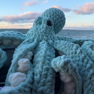 May include: A large, handmade, turquoise crochet octopus with white accents. The plush toy has a round head, large eyes, and eight textured tentacles. The background shows a cloudy sky and ocean.