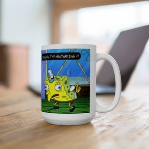 May include: White ceramic mug with a cartoon image of Spongebob Squarepants and the text "Do you try restarting it?". The mug has a large handle and is on a wooden surface.