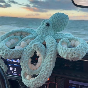 May include: A large, light blue crocheted octopus with white accents sits on a car dashboard. The plush toy has multiple tentacles and a round head. The background shows a beach and ocean under a cloudy sky.