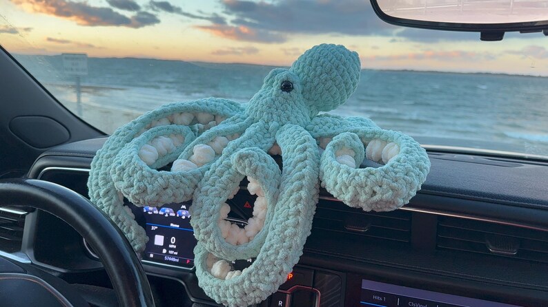 May include: A large, light blue crocheted octopus with white accents sits on a car dashboard. The plush toy has multiple tentacles and a round head. The background shows a beach and ocean under a cloudy sky.