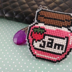 May include: A cross-stitch jam jar keyring. The pink jar has a brown lid and a red strawberry. The word "JAM" is stitched in white. The keyring is attached to a silver chain. The background includes a black quilted purse.