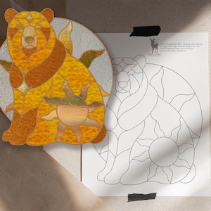 Op de afbeelding: Een cirkelvormig glas-in-loodpaneel met een zittende beer in amber-, goud- en bruintinten met een zonmotief, vergezeld van een bijpassend zwart-wit lijnpatroonblad met de tekst "Solar Celestial Sun Bear Stained Glass Pattern".