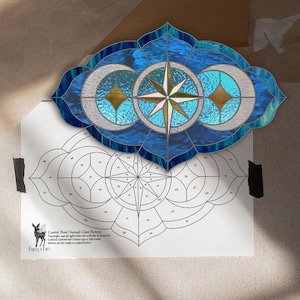 May include: A stained glass panel in shades of blue, featuring a central compass rose design, flanked by crescent moons and diamond shapes. The panel is mounted above a pattern diagram. The text "Cosmic Panel Stained Glass Pattern" is visible.