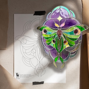 May include: A stained glass suncatcher shaped like a luna moth with vibrant green and purple glass segments, featuring dark red circular accents on the wings. Beside it is a white paper pattern sheet labelled Luna Moth Stained Glass Pattern.