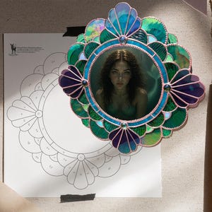 May include: A stained glass frame with a portrait of a woman. The frame is a mix of green, blue, and purple glass. The frame is round with a scalloped edge. The portrait is of a woman with long brown hair.