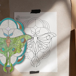 May include: A stained glass design featuring a green and brown moth with blue accents, set within a teal and white frame. A black and white line drawing of the moth is next to the stained glass piece.
