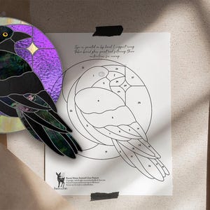 May include: A stained glass raven design with a purple and yellow background. The raven is black with iridescent details. A line drawing of the raven is next to the glass art. The text on the drawing says "Raven Moon stained glass patterns".