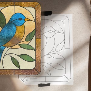 May include: Stained glass panel featuring a blue bird perched on a branch with green leaves. The bird has a textured appearance, and the background is a warm yellow. The panel is framed in a brown border. A line drawing of the bird is next to the panel.