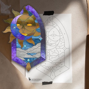 May include: A stained glass sun and moon design with a purple border. The sun has a face with yellow eyes and a golden hue, while the moon is a crescent shape. The design includes blue, white, and gold elements. A paper pattern is next to the stained glass.