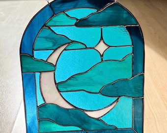 May include: An arched stained glass suncatcher featuring a white crescent moon and a small four-pointed star set against a background of layered clouds in various shades of blue and teal, held together by dark metal solder lines and a chain.