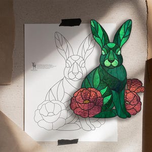 May include: A stained glass rabbit in shades of green with red flowers, displayed alongside a black and white line art pattern of the same rabbit and floral design on a white sheet of paper, featuring clear geometric segments and dark lead lines.