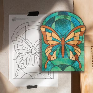 May include: A stained glass butterfly with orange and teal wings and a brown body. The butterfly is set against a teal and green background. A black and white line drawing of the butterfly is on a white sheet of paper.