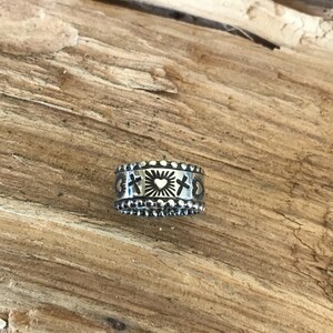 Op de afbeelding: Een zilverkleurige ring met een gedetailleerd ontwerp met kruisen, harten en zonnestralen. De ring heeft een kralenrand en is geplaatst tegen een houten achtergrond. De ring is een sieraad.