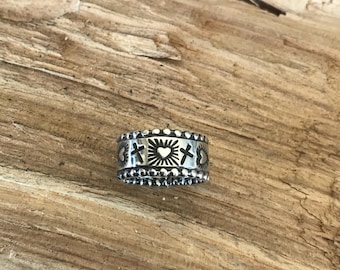Puede incluir: Un anillo plateado con un diseño detallado con cruces, corazones y rayos de sol. El anillo tiene un borde de cuentas y está sobre un fondo de madera. El anillo es una joya.