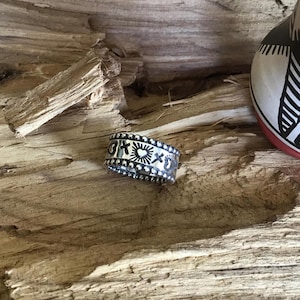 Peut inclure: Une bague en argent avec un motif détaillé comprenant des croix, des cœurs et des éclats de soleil. La bague est posée sur un fond en bois texturé, avec une partie d'un pot en céramique décoratif visible.
