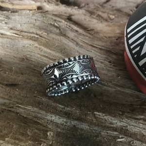 Op de afbeelding: Een zilveren ring met een parelrand en ingewikkelde geometrische ontwerpen. De ring ligt op een houten oppervlak.
