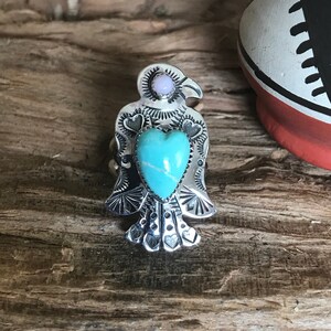 Op de afbeelding: Een zilverkleurige ring in de vorm van een vogel met een turquoise hartvormige steen. De vogel heeft een kleine, ronde, roze steen op de top van zijn kop. De ring ligt op een houten oppervlak.