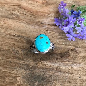 May include: A silver ring with a large, oval turquoise stone. The stone is a bright, vibrant blue with brown inclusions. The ring has a detailed, patterned band. The ring is set against a wooden background with purple flowers.
