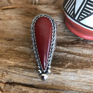 May include: A silver ring with a large, teardrop-shaped, polished red stone. The stone is set in a detailed silver band with a braided design around the edges. Small silver beads adorn the bottom of the stone. The ring is displayed on a wooden surface.